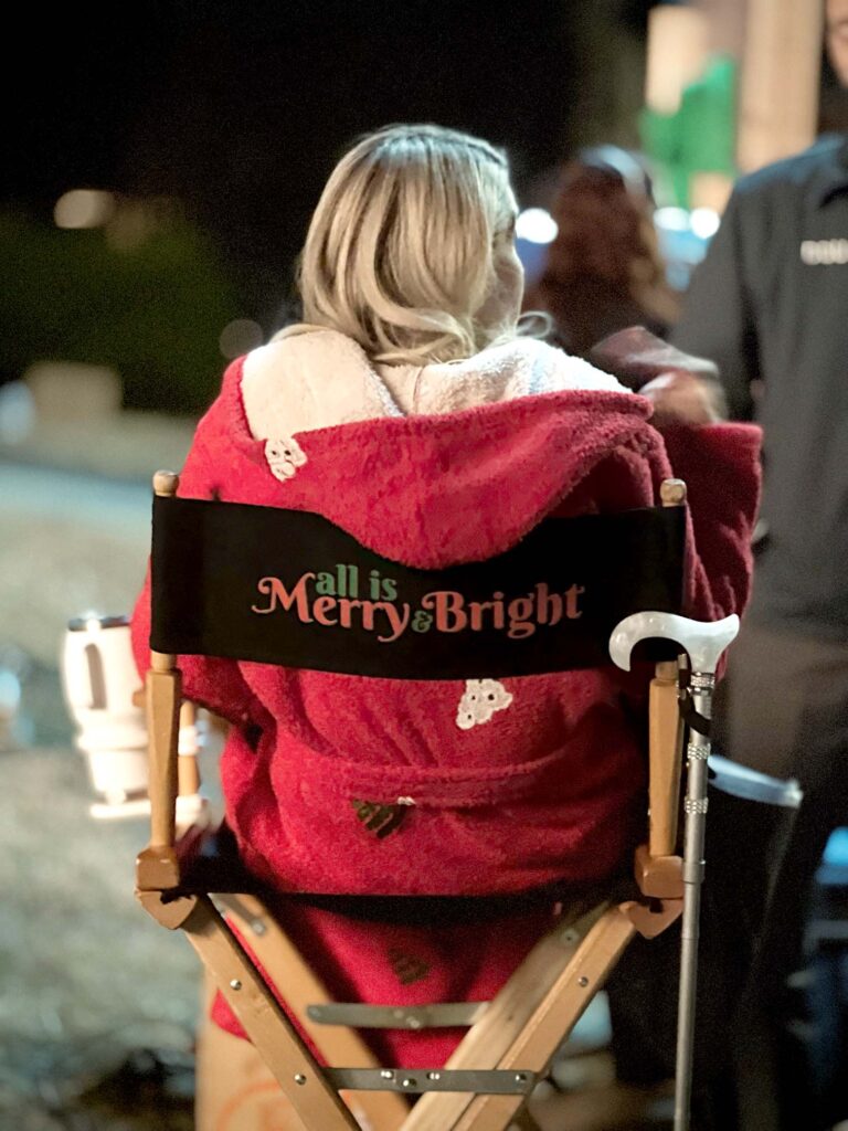 A person with blonde hair wearing a red robe sits in a director's chair with "All is Merry & Bright" printed on the backrest during a nighttime film shoot.
